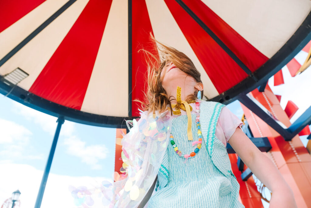 A child wearing a light blue dress and colorful necklace stands under a red-and-white striped canopy, wind blowing their hair across their face—a vibrant moment captured by a Brooklyn kids photographer on a bright, sunny day.
