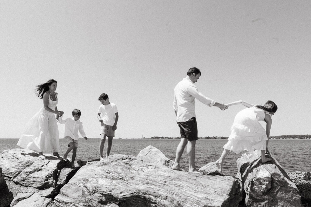 A family of five stands on large rocks by the water. Captured by a Brooklyn family photographer, the father holds a girl’s hand as she climbs, while the mother and two boys walk behind in light summer outfits under a clear sky.