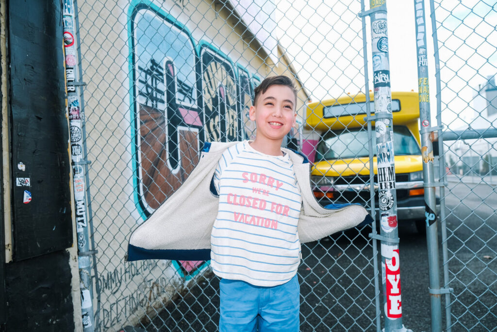 A smiling boy stands in front of a chain-link fence, spreading open his jacket. Captured by a Brooklyn kids photographer, he wears a striped shirt that reads SORRY, CLOSED FOR VACATION, with a yellow school bus and colorful graffiti behind him.