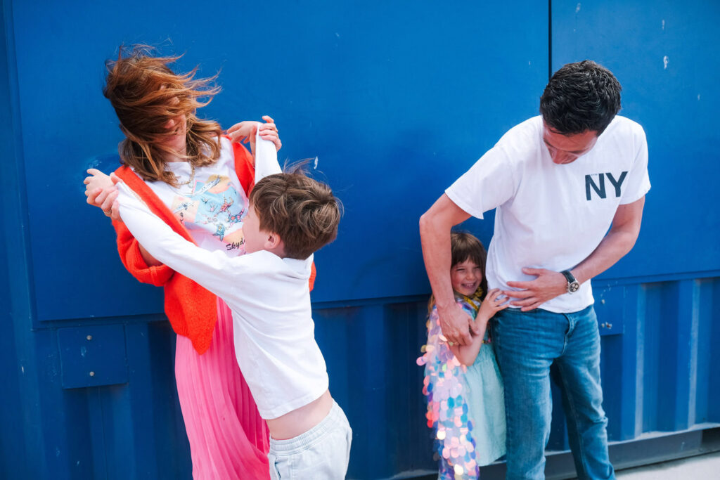 A woman and man playfully interact with two young children in front of a bright blue wall. Captured by a Brooklyn family photographer, the boy laughs as he swings arms with the woman, while the man tickles the girl, who smiles shyly and holds a shiny garland.