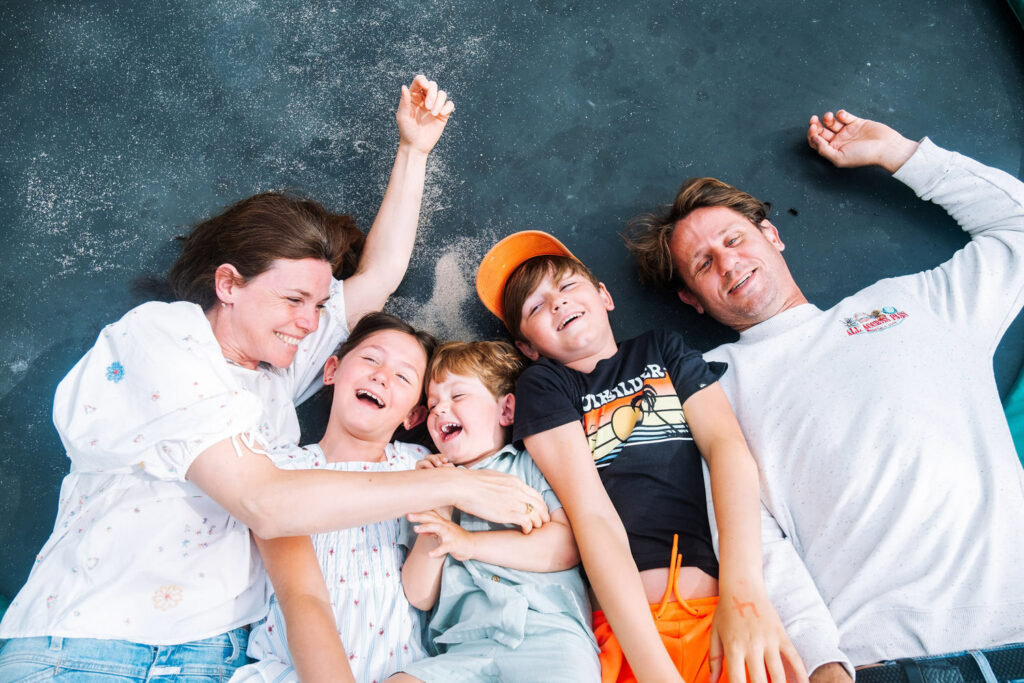 A family of five lies side by side on a dark surface, smiling and laughing together. The two adults are on either end, with three children in between—a joyful moment perfectly captured by a Brooklyn family photographer.