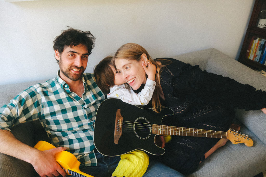 Family of three laughing together on couch with guitars - NYC family photography capturing authentic moments and storytelling