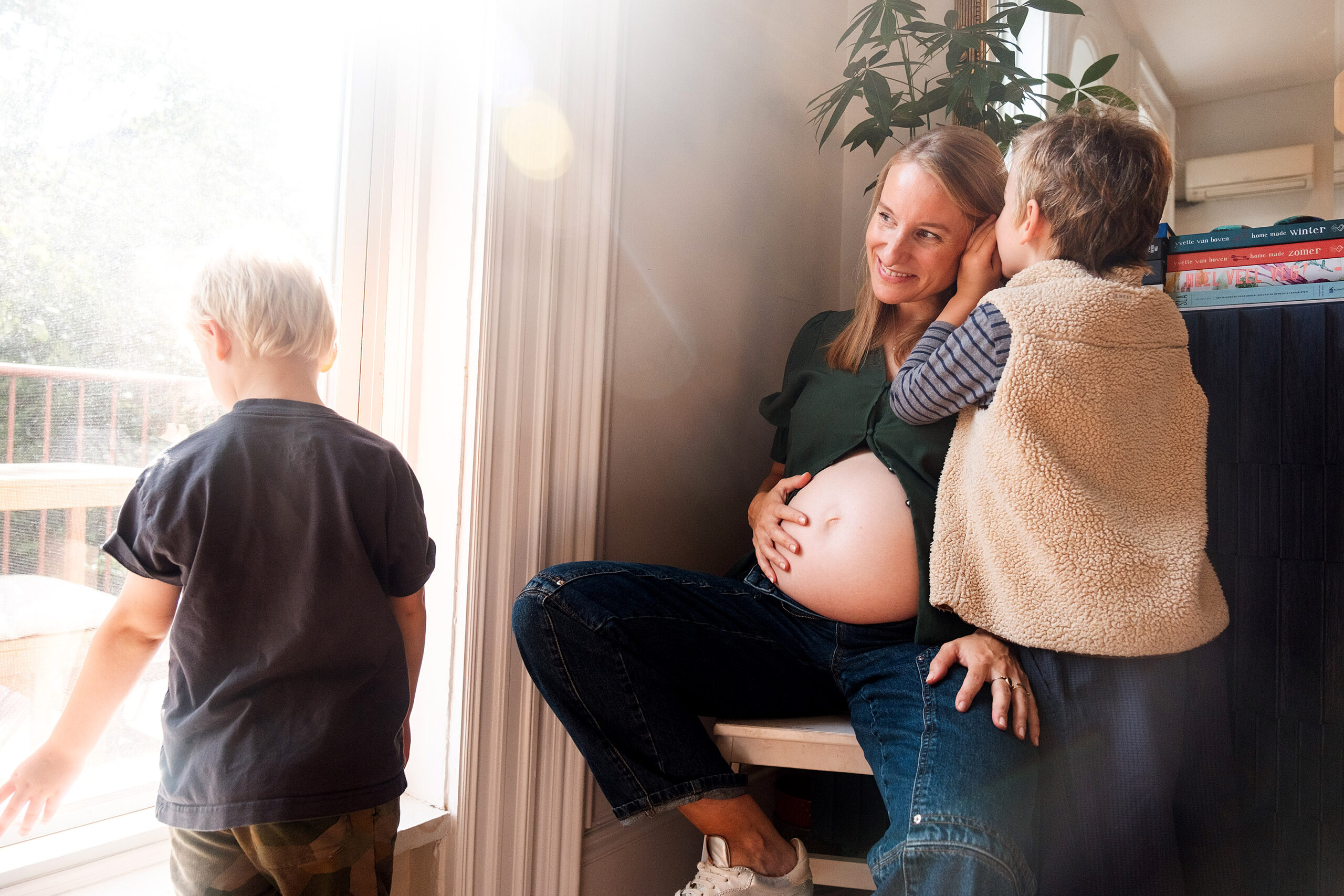Pregnant woman sitting by window with two children in Brooklyn home during maternity photography session
